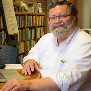 David Crumm sitting at a desk with a laptop. There are shelves filled with books in the background.