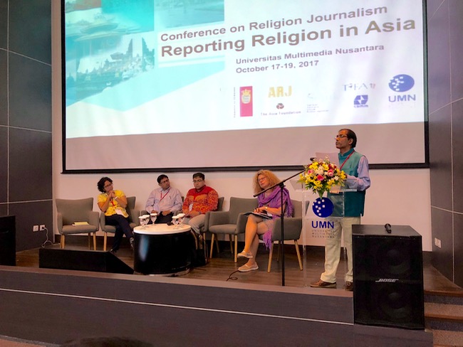 A man stands and speaks into a microphone on a stage with a slide projected behind him stating ”Conference on Religion Journalism; Reporting Religion in Asia”