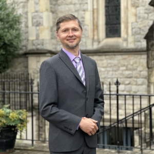 Professional photograph of Maksym Balaklytskyi in a gray suit and purple shirt and tie, standing in front of a stone church building.