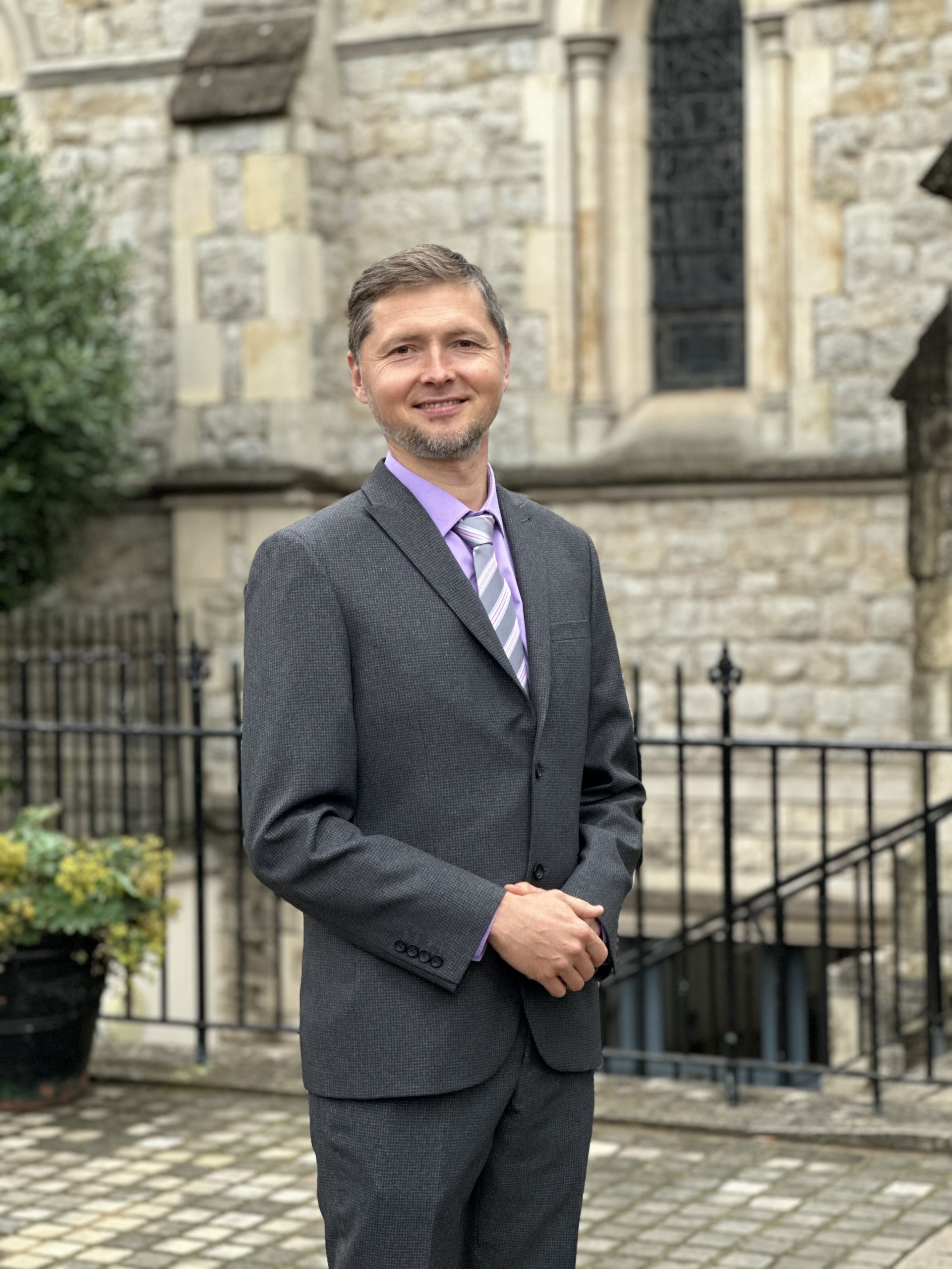 Professional photograph of Maksym Balaklytskyi in a gray suit and purple shirt and tie, standing in front of a stone church building.